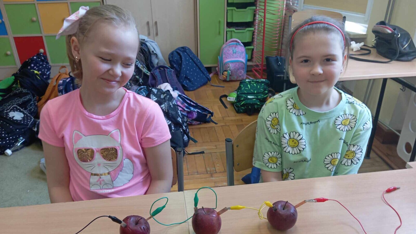 Dziewczynki siedzą przy stole, przed nimi stoi bateria zbudowana z jabłek.