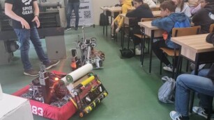 Uczniowie siedzą w ławkach i oglądają jak wykładowca POLLUB steruje skonstruowanym robotem.
