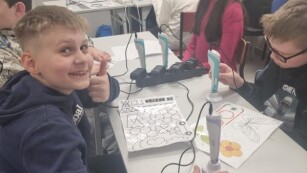 Uczniowie klasy 5c siedzą przy stole i przygotowują się do pracy długopisami 3D. Na pierwszym planie widzimy uśmiechającego się chłopca w granatowej bluzie.