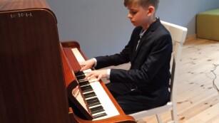 Przy fortepianie siedzi chłopiec, gra na instrumencie.