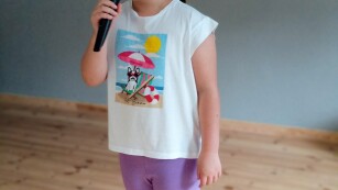 Dziewczynka stoi na scenie, trzyma w ręce mikrofon, śpiewa.
