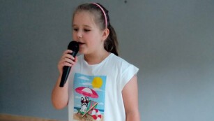 Dziewczynka stoi na scenie, trzyma w ręce mikrofon, śpiewa.