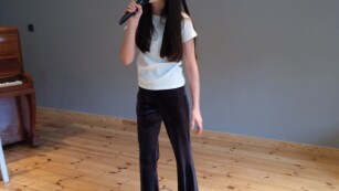 Dziewczynka stoi na scenie, trzyma w ręce mikrofon, uśmiecha się, śpiewa.