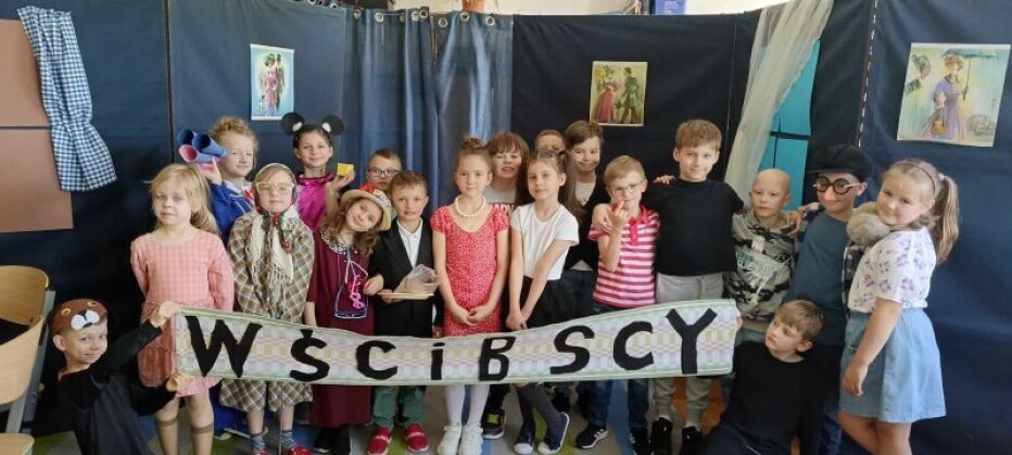 Grupa dziewiętnaściorga dzieci stoi trzymając napis „Wścibscy”.